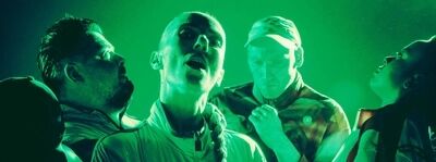 Beeld bij The Current: vier spelers staan dicht bij elkaar in fel groen licht en kijken omhoog. De middelste, met een vlecht over de schouder, heeft de mond open; de anderen kijken van de camera weg.