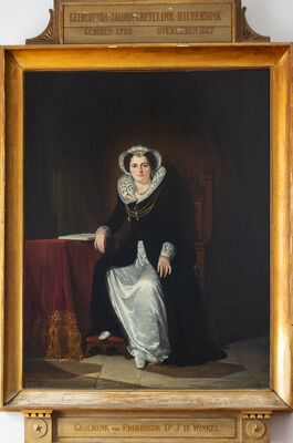 Geschilderd portret ten voeten uit van een vrouw in een zwarte japon met een witte kanten kap en molensteenkraag, staand naast een tafel met een rood kleed waarop een opengeslagen boek ligt. Op het plaatje boven de lijst: Geertruyda Jacoba Grevelink-Hilverdink, geboren 1786, overleden 1827; eronder: Geschenk van Professor Dr. J. te Winkel.