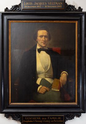Geschilderd portret van een zittende man in een zwarte jas met een wit vest en zwarte strik, een boek in zijn linkerhand. Op het plaatje boven de zwarte lijst: Louis Jacques Veltman, 29 December 1817 – 10 November 1907; eronder: Geschenk der familie.