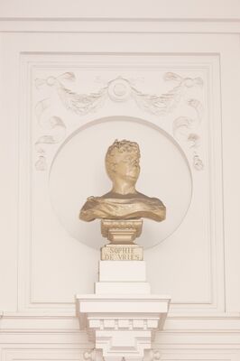 Bronskleurige buste van een vrouw met opgestoken haar, kijkend omhoog en opzij, op een sokkel met het opschrift SOPHIE DE VRIES, in een ronde nis in een wit gestucte wand met een guirlande erboven.