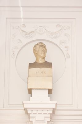 Roomwitte gipsen buste van een man met krullend haar, het hoofd geheven en licht opzij gedraaid, op een sokkel met het opschrift SNOEK, in een ronde nis in een wit gestucte wand met een guirlande met linten erboven.
