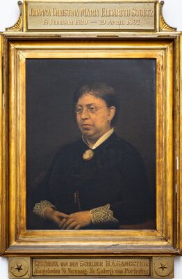 Geschilderd portret van een vrouw met een lorgnet, in een zwarte japon met een witte kraag, een cameebroche en brede kanten manchetten; haar handen liggen gevouwen in haar schoot. Op het plaatje boven de lijst: Johanna Christina Maria Elisabeth Stoetz, 18 Februari 1829 – 19 April 1897; eronder: Geschenk van den schilder H.A. Sangster.