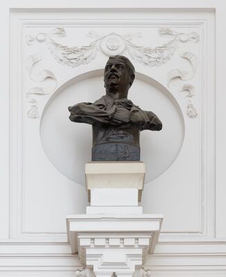 Bronzen buste van een man met een snor, het hoofd opgeheven en de mond half geopend, in een hemd met een losse strik; op de voet een reliëf van een figuur met een harp aan het water. De buste staat in een witte nis onder een pleisterguirlande.