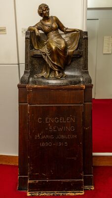 Bronzen beeld van een zittende vrouw in een lange geplooide jurk op een donkere stenen bank, met één arm over de rugleuning. Het geheel staat op een houten sokkel met de ingesneden tekst C. ENGELEN-SEWING, 25 JARIG JUBILEUM 1890-1915, en de signatuur FRANS WERNER FEC.
