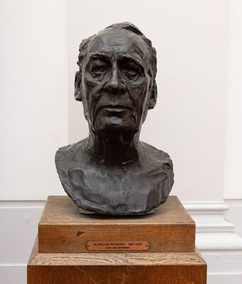 Bronzen buste van een man met een smal, ingevallen gezicht en achterovergekamd haar, op een eiken sokkel. Op het plaatje: Dr. Willem Royaards 1867–1929, door Mari Andriessen.
