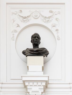 Bronzen buste van een oudere man met een streng, hoekig gezicht en een mantel over de schouders, opkijkend. De buste staat op een lichte sokkel in een witte nis onder een pleisterguirlande.