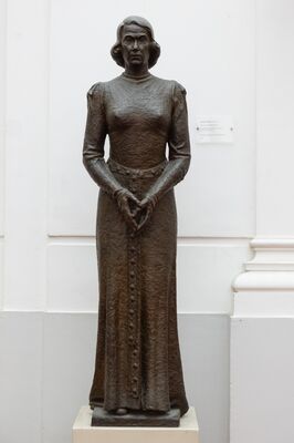 Bronzen staand beeld ten voeten uit van een vrouw in een lange jurk met een rij knopen over de volle lengte, de handen losjes voor zich gevouwen, op een stenen voetplaat op een lichte sokkel. Rechts aan de wand een klein informatiebordje.