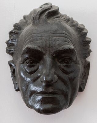 Donkergroen bronzen gezichtsmasker aan een witte wand: een oudere man met achterovergekamd, warrig haar, diepliggende ogen onder zware wenkbrauwen en een naar beneden getrokken mond.