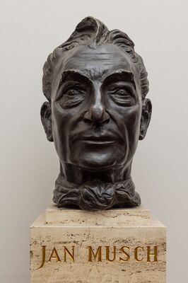 Bronzen kop van een oudere man met achterovergekamd haar, diepe lachrimpels en een losgeknoopte das, op een travertijnen sokkel met de vergulde naam JAN MUSCH.