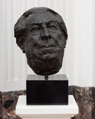Bronzen kop van een oudere man met een grof bewerkt oppervlak, zware wenkbrauwen en achterovervallend haar, op een zwarte sokkel.