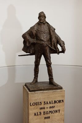 Bronzen beeldje ten voeten uit van een man met een baret en een wijde mantel, breeduit staand met een degen aan zijn zij. Op de travertijnen sokkel: Louis Saalborn 1891 – 1957, als Egmont, 1932.