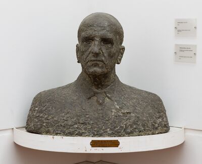 Donkere bronzen buste van een kale man in colbert en stropdas, met een strenge blik recht vooruit, op een witte hoekplank met een messing plaatje: Mr A. de Roos, wethouder van Amsterdam voor het onderwijs en de kunstzaken van 1945 tot 1962.