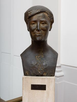 Bronzen buste van een glimlachende vrouw met een strakke muts of badkap, met ruw bewerkte schouders, op een travertijnen sokkel met het plaatje: Netty Hamel.