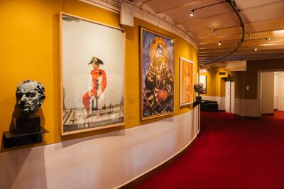 Foyergang met okergele wanden en rood tapijt tijdens de Openbare rondleiding met het Amsterdam Museum, met aan de wand een portretfoto van een acteur in harlekijnkostuum en twee bronzen hoofden op sokkels.