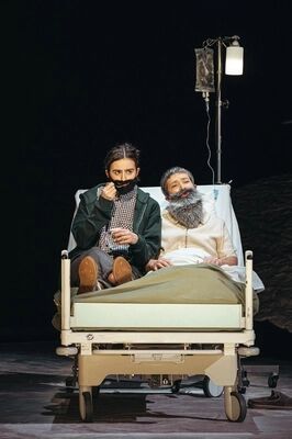 Scène uit No Yogurt for the Dead - Histoire(s) du Théâtre VI: twee spelers met valse baarden zitten samen op een ziekenhuisbed. De een houdt een bekertje vast en drukt een zwarte baard tegen het gezicht, de ander leunt achterover met een grijze baard. Rechts staat een infuusstandaard met lamp.