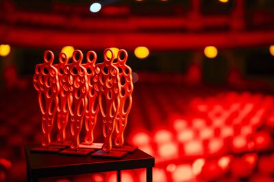 Beeld bij Winq Awards 2026: zes rode beeldjes in de vorm van menselijke figuren met ronde uitsparingen staan naast elkaar op een tafeltje. Op de achtergrond ligt, onscherp, de rood verlichte theaterzaal met balkons.