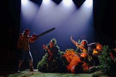 Scène uit Oh oh oh dennenboom (6+): een speler met rode muts richt een grote kettingzaag op drie spelers die als kerstbomen zijn uitgedost en hun handen afwerend opsteken. Rechts hangt een label met het woord 'ONGESCHIKT'.