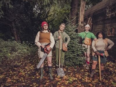 Campagnebeeld voor Oh oh oh dennenboom (6+): vier spelers staan in een herfstig bos naast een kerstboom. Links een vrouw met rode muts en kettingzaag, daarnaast een oudere man met een schep, dan een man in een groene trui met fototas, en rechts een vrouw met een bijl.