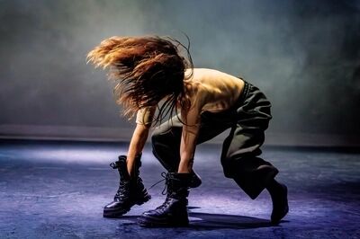 Scène uit SEXODUS: een danseres buigt diep voorover met het haar over haar gezicht, in een wijde donkergroene broek en zware zwarte laarzen, op een blauw verlichte vloer.
