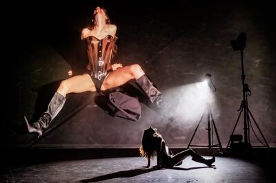 Scène uit SEXODUS: een danseres springt met gespreide benen door de lucht in een glanzend korset en hoge laarzen; op de vloer eronder buigt een tweede danseres achterover. Rechts staan filmlampen op statieven.