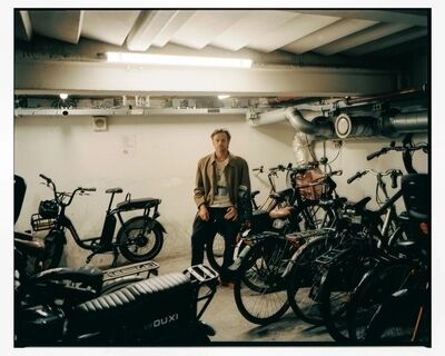 Sanne den Hartogh staat in een ondergrondse stalling tussen geparkeerde fietsen en scooters, in een beige jas.