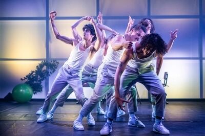 Scène uit Controle: vijf spelers in witte hemden, lichte ribfluwelen broeken en witte sneakers bewegen voorovergebogen en met opgeheven handen in dezelfde richting. Achter hen staat een wand van lichtgevende panelen, met een groene gymbal en de schaduw van een plant.