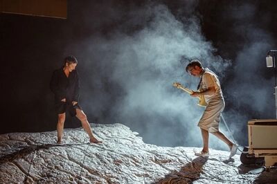 Scène uit No Yogurt for the Dead - Histoire(s) du Théâtre VI: twee spelers lopen door de rook over een zilvergrijs landschap. Links een speler in een zwart jasje met blote benen, rechts iemand in een beige jurk die een basgitaar bespeelt; uiterst rechts staat een ziekenhuisbed.