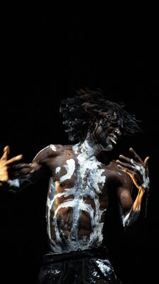 Beeld bij As we are: een danser met ontbloot bovenlijf, besmeurd met witte verf over borst, hals en gezicht, danst met wapperende dreadlocks en wijd gespreide armen in een donkere ruimte.