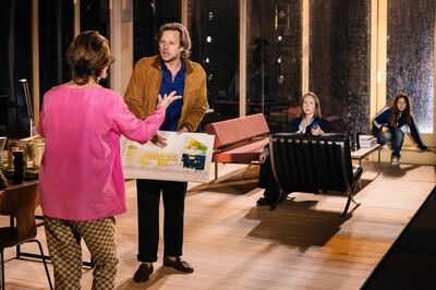 Sanne den Hartogh staat in een lichte woonkamer op het toneel en gebaart naar een speelster in een roze trui die hem een maquette voorhoudt; op de achtergrond kijken twee anderen toe.