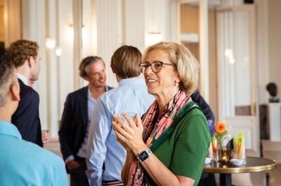 Met een glas in beide handen glimlacht een vrouw met kort blond haar en een bril, in een groen shirt met een roze gedessineerde sjaal; achter haar praten mannen in overhemden en colberts, en rechts staat een statafel met een oranje bloem en groentesticks.