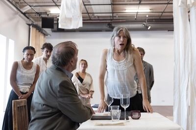 Een man zit aan tafel en een vrouw staat bij hem aan tafel met een verraste blik. Op de achtergrond staat een groep mensen te kijken.