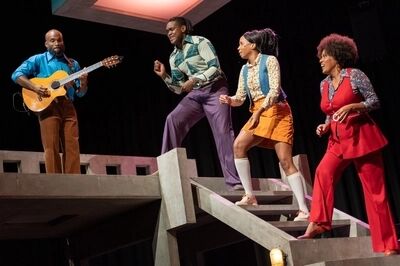 Scène uit De Suriname-trilogie: De Gliphoeve: vier spelers op een decor van betonnen trappen en galerijen. Links speelt een man in een blauw overhemd gitaar, de andere drie bewegen dansend over de trap in kleding uit de jaren zeventig.