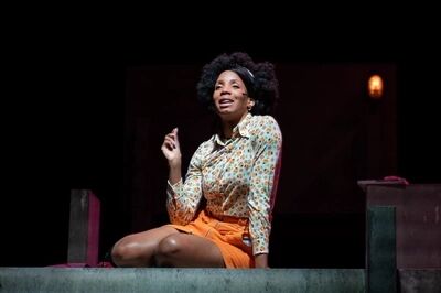 Scène uit De Suriname-trilogie: De Gliphoeve: een speelster met afro en haarband, in een gebloemde blouse en een oranje rok, zit op een betonnen rand en heft haar wijsvinger terwijl ze spreekt.