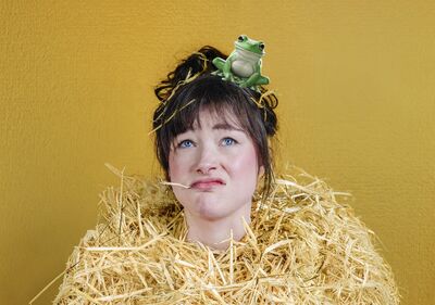 Campagnebeeld voor Meiden & Prinsen: een vrouw met opgestoken haar en een strootje in haar mond kijkt omhoog. Ze draagt een pak van stro en op haar hoofd zit een groene kikker; de achtergrond is okergeel.