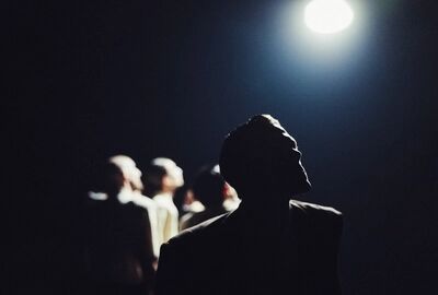 Beeld bij GONE - Inspired by Benjamin Clementine: een danser staat met de rug naar de camera in het donker en kijkt omhoog naar één felle lamp. Achter hem staan onscherpe figuren in het halfduister.