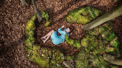 Beeld bij Grondtonen (9+): van bovenaf gezien ligt een kind in een lichtblauwe jurk op de bosbodem, tussen bruine beukenbladeren en plukken groen mos, met blote voeten en een arm omhoog. Rondom staan boomstammen.