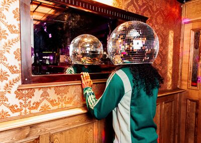 Campagnebeeld voor EN NOU GAAN WE EVEN HELEMAAL UIT ONZE PLAAT (een topshow) (6+): een speler met krullend haar en een grote discobol over het hoofd staat in een groen trainingsjack voor een spiegel in een zaal met goudkleurig behang; in de spiegel is de discobol weerspiegeld.