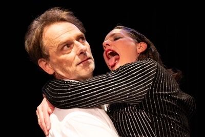Scène uit Oedipus: een speler in een wit overhemd staat in het donker terwijl een speelster in een glinsterend gestreept jasje haar arm om zijn hals slaat en met open mond en uitgestoken tong lacht.