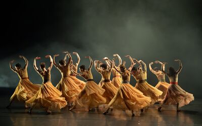 Beeld bij Nederlands Balletgala: een groep dansers in okergele jurken met wijd uitwaaierende rokken draait over het podium, de armen gebogen boven het hoofd. De achtergrond is donker en rokerig.