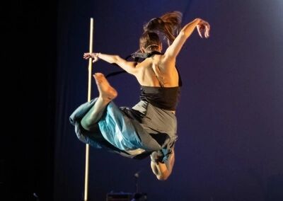 Beeld bij Negen: een danseres springt met opgetrokken benen door de lucht, met de rug naar de camera en de armen gespreid. Links staat een gele verticale staaf; de achtergrond is donkerblauw.