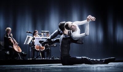 Twee dansers in A poem for the world: een danser in het zwart zit op de grond met gestrekte benen en houdt een danseres in het wit vast, die zich achterover over hem heen buigt met de armen boven het hoofd. Op de achtergrond spelen twee cellisten en een musicus achter een lessenaar.