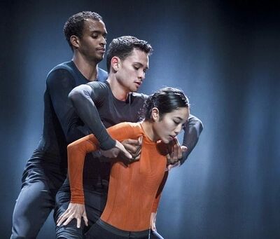 Beeld bij A poem for the world: drie dansers staan dicht tegen elkaar in blauw licht. Vooraan buigt een danseres in een oranje shirt haar hoofd naar beneden; achter haar staan twee dansers in donkere shirts, van wie er een zijn arm om haar schouder legt.