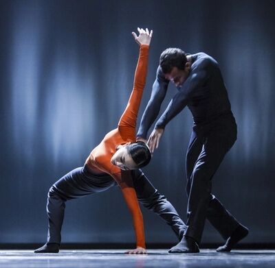 Beeld bij A poem for the world: een danseres in een oranje shirt staat wijdbeens en buigt opzij, met één arm hoog gestrekt en de andere hand op de vloer. Een danser in een blauwgrijs shirt buigt zich van achteren over haar heen en raakt haar arm aan.