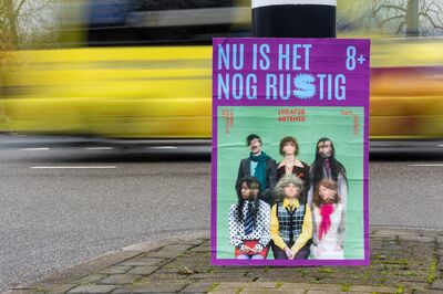 Campagnebeeld voor Nu is het nog rustig (8+): een affiche geplakt op een paal langs de weg, met in blauwe letters op paars 'NU IS HET NOG RUSTIG 8+' en daaronder zes spelers in bonte kleding voor een groene achtergrond. Op de weg erachter rijdt een gele bus voorbij.