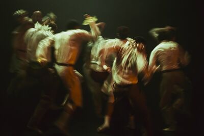 Beeld bij Out of the stillness: een groep dansers in lichte overhemden beweegt door een donkere ruimte; door de lange sluitertijd zijn de lichamen vervaagd tot vegen licht.
