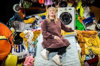 Portret bij Waste me (13+): een vrouw met blond haar in een bruine jurk en zwarte legging zit op een matras te midden van bergen kleding, tassen en een wasmachine, en kijkt in de camera.