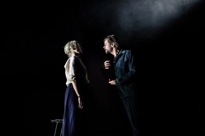 Op een verder zwart toneel staan in Prophet Song een vrouw met opgestoken blond haar in een witte blouse en een man met baard in een donker ribpak dicht bij elkaar in een bundel licht; hij praat tegen haar met zijn handen voor zijn borst.