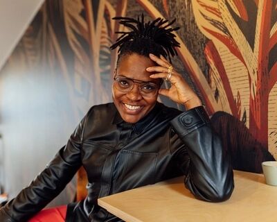 Portret bij Een groter verhaal: een vrouw met korte dreadlocks en een bril, in een zwarte leren blouse, zit aan een licht houten tafel met haar hand tegen haar slaap. Ze lacht naar de camera; achter haar een muurschildering met bladeren in rood, oker en zwart.