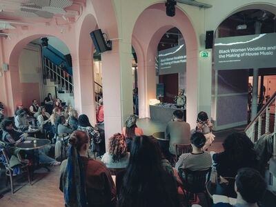 Beeld bij Rooted in Culture: UK Sounds influencing House culture: publiek zit aan cafétafels in een zaal met roze geschilderde bogen en kijkt naar een spreker achter een dj-tafel. Op twee schermen staat 'Black Women Vocalists and the Making of House Music', met daaronder 'Follow the work @lynneedenise'.