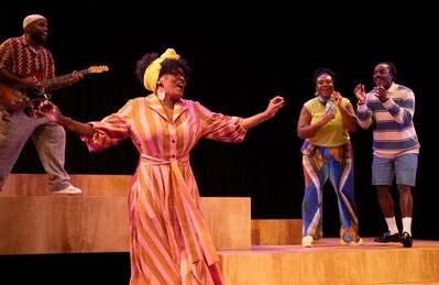 Scène uit De Suriname-trilogie: Stepping Stones: een speelster in een roze-oranje gestreepte jurk met gele hoofddoek zingt met wijd gespreide armen. Links speelt een muzikant gitaar op een houten podiumblok, rechts staan twee spelers te klappen.